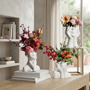 May include: Three white ceramic vases shaped like human heads with hands resting near the face. Each vase holds a different arrangement of colorful artificial flowers, including red berries, pink petals, and green foliage, displayed on white books.