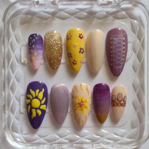 May include: A set of ten almond-shaped press-on nails in a clear plastic case. The designs include purple and yellow gradients, gold glitter, yellow flowers on a yellow base, solid cream, purple corset lacing, a yellow sun, shimmer, and a crown.