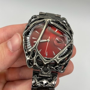 Op de afbeelding: Een polshorloge met een dieprode wijzerplaat en zilverkleurige uurmarkeringen en wijzers. De horlogekast en de schakelarmband zijn omhuld door een donker, staalgrijs metalen frame met een complex, organisch, web- of takachtig uitgesneden ontwerp.