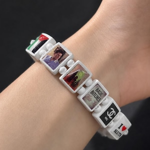 Op de afbeelding: Een witte kunststof schakelarmband met rechthoekige bedels met diverse prints, waaronder een vlag, een persoon, een groene afbeelding, de tekst BASIC BITCHES Hate Me, een logo en de tekst I heart SLUTS met een rood hartje.