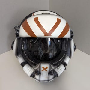 Puede incluir: Un casco blanco y negro con una visera tintada oscura y un gráfico en forma de V de color marrón en la frente. La parte frontal incluye un símbolo X marrón central, detalles de ventilación laterales y un acabado desgastado.