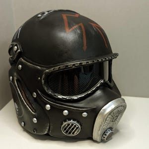 Op de afbeelding: Een matzwarte tactische helm met een verweerde zilveren afwerking op de randen en klinknagels. De helm is voorzien van een donker gaasvizier, een opvallend ademhalingsfilter aan de voorzijde en een rood bliksemsymbool op de bovenkant.