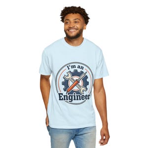 May include: A light blue short-sleeve t-shirt featuring a circular graphic on the chest. The design includes a dark blue gear, a crossed wrench and soldering iron, and the text "I'm an Engineer" in bold dark blue lettering.