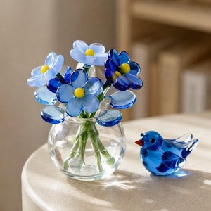 Op de afbeelding: Een bolvormige vaas van helder glas met een boeket handgemaakte glazen bloemen in lichtblauw, donkerblauw en met gele harten, vergezeld door een klein, massief blauw glazen vogelbeeldje met een piepkleine oranje snavel en zwarte oogjes.