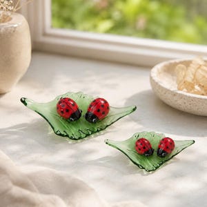Könnte beinhalten: Zwei grüne Glasschalen in Blattform, die jeweils zwei kleine rote Marienkäfer-Figuren mit schwarzen Punkten und schwarzem Kopf enthalten. Die Glasblätter haben eine texturierte, geäderte Oberfläche und geschwungene Ränder für eine Dekoration.