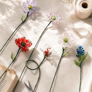 May include: A collection of six handcrafted glass flowers on thin green wire stems. The set includes a purple, a light pink, a vibrant red, a clear white, a second clear white, and a small blue cluster flower, each featuring delicate green glass leaves.