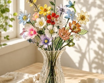 May include: A clear, ribbed glass vase holds a bouquet of multi-colored glass flowers. The arrangement features various blooms in shades of yellow, red, blue, pink, white, and orange, each with green glass stems and leaves, creating a vibrant display.