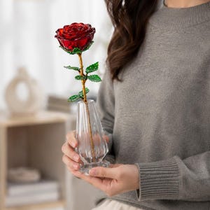 May include: A decorative artificial rose with deep red petals, a gold-toned stem, and green crystal-encrusted leaves. The flower is displayed in a clear, faceted glass vase with a tapered shape and a wide, stable base.