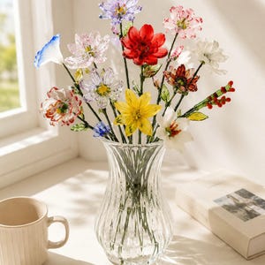 May include: A clear glass vase containing a bouquet of multi-colored glass flowers. The arrangement features various blooms in shades of red, yellow, white, pink, blue, and purple, each with green glass stems and leaves, displayed in a fluted glass vessel.