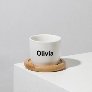 May include: A white ceramic cup with the name Olivia printed in black sans-serif font on the front. The cup rests on a circular, light brown wooden coaster with a smooth, rounded edge, creating a simple and functional drinkware set.