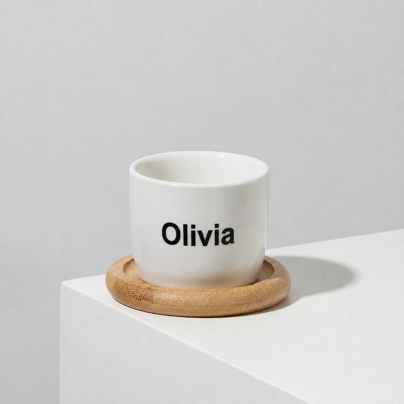 May include: A white ceramic cup with the name Olivia printed in black sans-serif font on the front. The cup rests on a circular, light brown wooden coaster with a smooth, rounded edge, creating a simple and functional drinkware set.