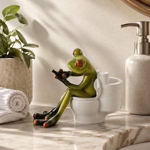 Op de afbeelding: Een groen kikkerbeeldje met grote rode ogen zittend op een wit keramisch toilet. De kikker houdt een klein zwart rechthoekig voorwerp in de handen. De toiletvoet is voorzien van een kleine cilindrische houder voor een rol toiletpapier.