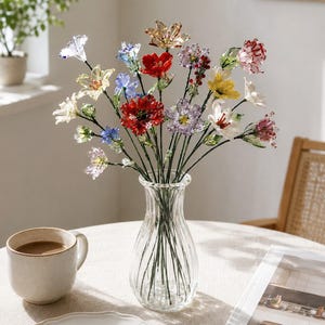 May include: A clear glass vase containing a bouquet of multi-colored glass flowers. The arrangement features individual stems with petals in shades of red, blue, yellow, purple, white, and pink, each attached to thin green wire stems.
