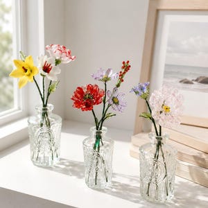 May include: Three clear glass bottles with embossed circular designs, each containing a small arrangement of colorful glass flowers. The flowers feature various petals in shades of yellow, white, red, purple, and pink, supported by thin green stems.