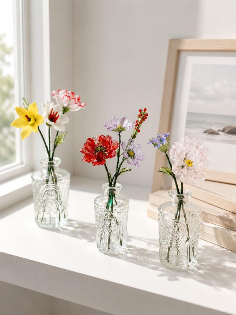 May include: Three clear glass bottles with embossed circular designs, each containing a small arrangement of colorful glass flowers. The flowers feature various petals in shades of yellow, white, red, purple, and pink, supported by thin green stems.