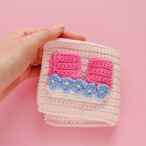 May include: A pink crocheted cup cosy with pink and blue flower embellishments.