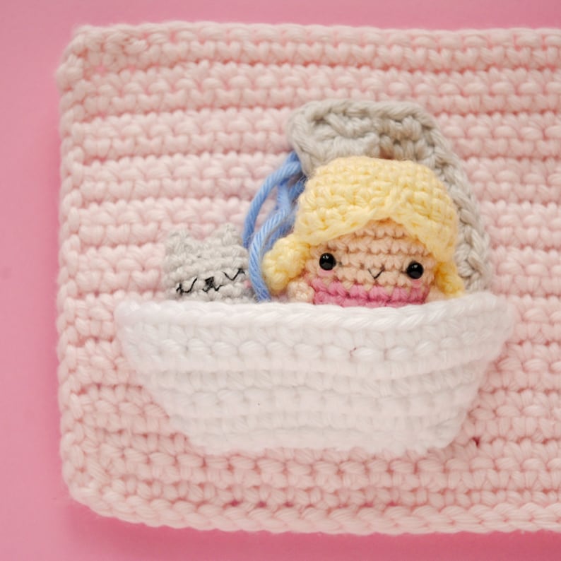 May include: A crocheted doll with yellow hair and a pink dress is sitting in a white crocheted bathtub. A grey crocheted cat is peeking out from behind the doll.