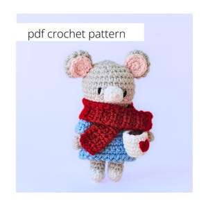 May include: A crocheted mouse wearing a red scarf and blue dress, holding a white mug with a red heart. The mouse has gray fur, pink ears, and a white nose. The text "pdf crochet pattern" is in the upper left corner.