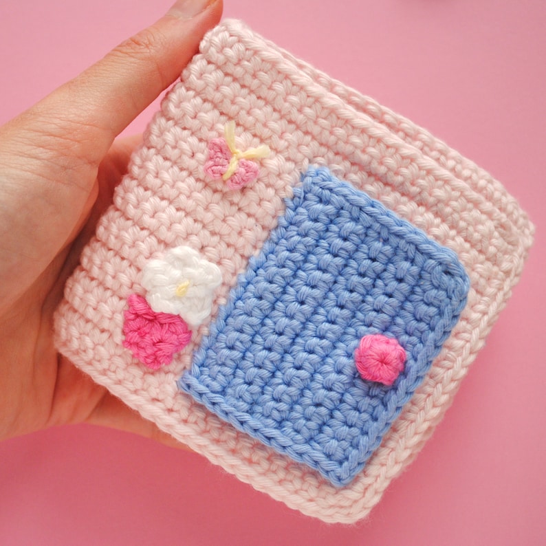 May include: A pink crocheted pouch with a blue crocheted pocket and pink and white flower embellishments.