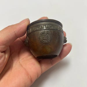 Könnte beinhalten: Ein kleines Gefäß aus dunkel bronzefarbenem Metall mit bauchigem Körper und weiter Öffnung. Die Außenseite zeigt ein dekoratives Band mit wiederkehrenden Schnörkelmustern am Rand und ein kreisförmiges Emblem mit einem Relief in der Mitte.