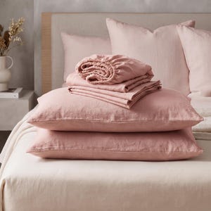 May include: A set of dusty rose bedding featuring two rectangular pillows stacked horizontally, topped with a folded fitted sheet and a flat sheet. The fabric has a visible woven texture, and the set is arranged on a neutral-coloured bed.