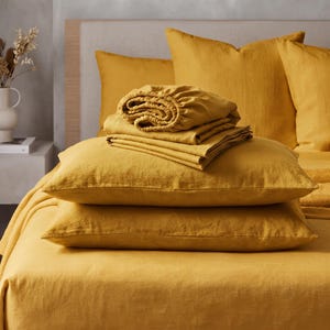 May include: A set of mustard yellow bedding featuring a fitted sheet, a flat sheet, and two pillowcases. The items are stacked neatly on a bed, showcasing the textured fabric. The set includes square pillow shams and a matching bed sheet.