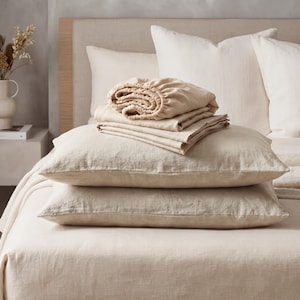 May include: A set of beige linen bedding including two rectangular pillows stacked on a flat sheet, with a folded fitted sheet placed on top. The fabric has a visible woven texture, and the items are arranged on a bed with a light-colored headboard.