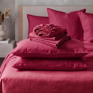 May include: A set of deep magenta linen bedding including a fitted sheet, a flat sheet, and two pillowcases. The items are stacked neatly on a bed, featuring a textured fabric finish and a consistent, rich, solid red-pink colour throughout.