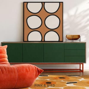 May include: A square framed wall art piece featuring a grid of six off-white circles outlined in black against a solid terracotta background. The artwork is displayed on a dark green sideboard with orange legs, positioned near an orange chair.