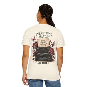 May include: A cream-colored t-shirt featuring a graphic on the back of a black vintage typewriter surrounded by pink roses and butterflies. The text reads: EVERYTHING CHANGED. new chapter different me. SO DID I. The design includes small star accents.