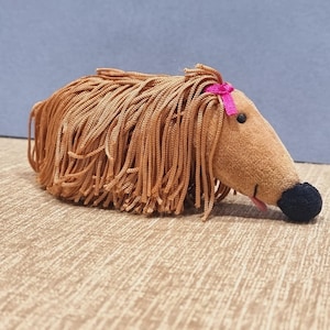 May include: A plush toy shaped like a creature with a tan velvet body, a black nose, and a small pink tongue. The toy features long, light brown yarn strands covering its back and sides, accented by a small, bright pink ribbon on its head.