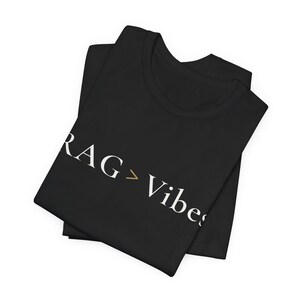 May include: A black t-shirt featuring the white text RAG > Vibes printed across the front. The greater than symbol is coloured in a muted gold tone. The shirt is folded, displaying a crew neckline and short sleeves in a simple, clean layout.