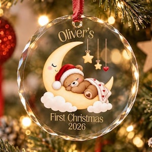 Könnte beinhalten: Ein runder, klarer, facettierter Weihnachtsbaumschmuck aus Glas mit einer Illustration eines braunen Teddybären mit roter Weihnachtsmütze, der auf einem gelben Halbmond mit weißen Wolken, Sternen und dem Text Oliver's First Christmas 2026 schläft.