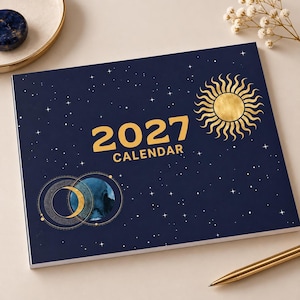 Könnte beinhalten: Ein rechteckiger Kalender für 2027 mit dunkelblauem Einband, verziert mit einer goldenen Sonnengrafik, einer Mondsichel und weißen Sternen. Der Text 2027 CALENDAR ist in großen goldenen Buchstaben mittig auf dem Cover aufgedruckt.