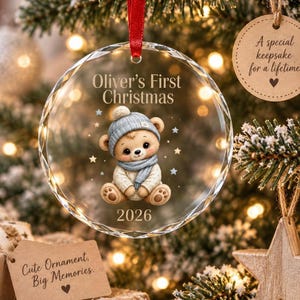 Könnte beinhalten: Ein runder, klarer Glas-Weihnachtsanhänger mit einer Illustration eines Teddybären, der eine blaue Strickmütze und einen Schal trägt. Der Text lautet Oliver's First Christmas und 2026. Der Anhänger hängt an einem roten Band und hat facettierte Kanten.