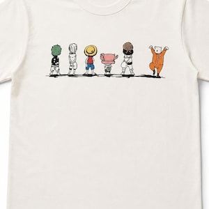 May include: A cream-coloured short-sleeve t-shirt featuring a graphic print of six cartoon characters standing in a row, viewed from behind. The characters include figures with green hair, a straw hat, a pink hat, and an orange jumpsuit.