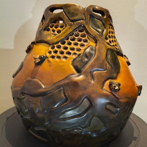 May include: A gourd vessel featuring a carved honeycomb pattern and raised bee figures. The surface displays a gradient of warm orange and deep brown tones, with organic, flowing dark brown shapes integrated into the design around the honeycomb sections.