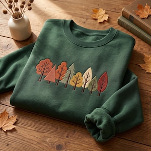 May include: A forest green crewneck sweatshirt featuring an embroidered row of stylized trees in autumn colors including orange, yellow, olive green, and brown. The design is centered on the chest, showcasing various leaf shapes and evergreen silhouettes.
