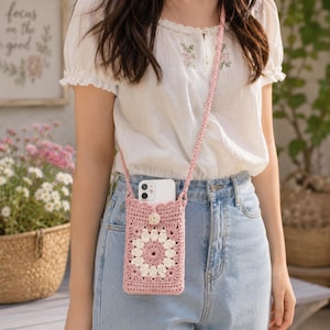 May include: A dusty pink crochet crossbody phone bag featuring a central white floral granny square motif, a matching long strap, and a single button closure at the top, currently holding a white smartphone inside the rectangular pouch.