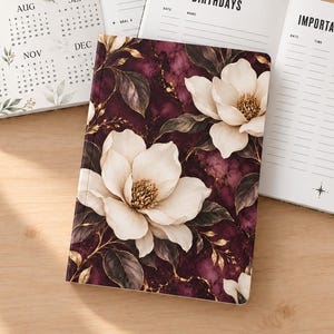 May include: Two open planners with white pages and a closed planner featuring a dark burgundy marble background with white magnolia flowers and gold accents. The open pages display a 2027 calendar, My Goals 2027, Birthdays, and Important Appointments.