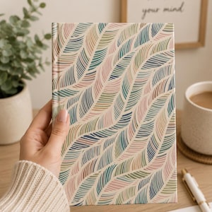 May include: A hardcover notebook featuring a repeating pattern of stylized, curved leaf shapes in muted shades of sage green, dusty pink, and navy blue against a cream-colored background. The design covers the entire front surface of the book.