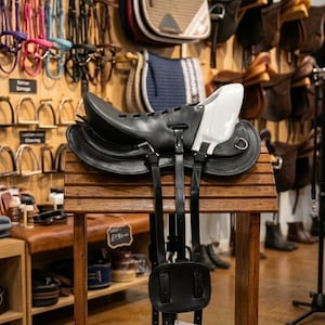 Pode incluir: Uma sela de cavalo em couro preto com um pomo branco, um assento contornado e longas tiras de couro preto penduradas verticalmente. A sela está montada num suporte de madeira, exibindo o seu design estrutural e componentes metálicos.