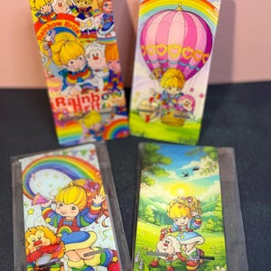 May include: Four rectangular metal phone stands featuring colorful illustrations of Rainbow Brite and her friends. Two stands are upright, displaying characters and a hot air balloon, while two are in clear plastic sleeves showing outdoor scenes.