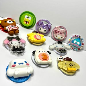 Könnte beinhalten: Eine Sammlung von zehn runden Handyhaltern mit verschiedenen Sanrio-Charakteren, darunter My Melody, Cinnamoroll, Pompompurin, Kuromi und Keroppi. Jeder Halter zeigt ein einzigartiges farbenfrohes Design, teilweise mit Strasssteinen oder 3D-geformten Elementen.