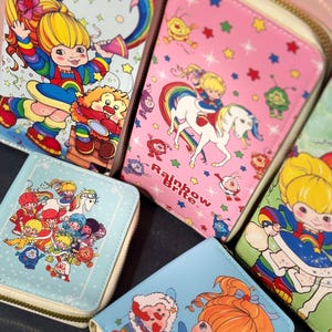 May include: A collection of four rectangular wallets featuring colorful illustrations of Rainbow Brite characters, rainbows, stars, and a white horse. The wallets have zip-around closures and light-colored trim, with one wallet clearly displaying the text Rainbow Brite.