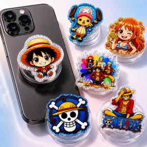 Pode incluir: Um smartphone preto com um suporte circular para telemóvel com uma personagem de desenho animado com um chapéu de palha. Cinco suportes circulares adicionais com vários designs de personagens de anime e um logótipo de caveira estão dispostos.