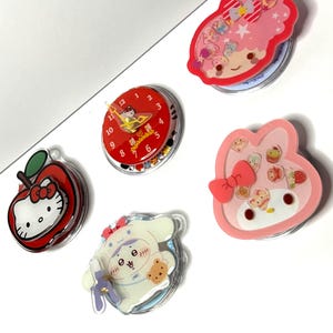 May include: Five circular decorative items featuring Sanrio characters. One is a red clock with a Hello Kitty face. Others include a red apple-shaped Hello Kitty, a pink rabbit-shaped My Melody with cupcakes, a white Cinnamoroll, and a pink Little Twin Stars.