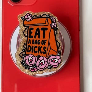 May include: A circular phone grip featuring a glittery gold border and a central orange paper bag illustration. The bag has the black text EAT A BAG OF DICKS printed on it, surrounded by pink roses with green leaves and black vine outlines.