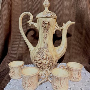 May include: A cream-coloured ceramic coffee pot with a matching lid and three small mugs. The set features intricate relief carvings of human faces and woodland scenes with a weathered finish. The pot has a tall handle and a curved spout.