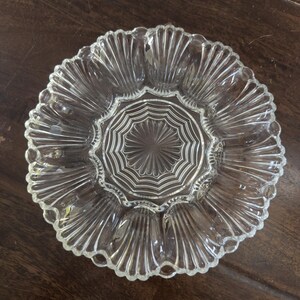 May include: A clear glass serving plate featuring a central circular medallion with a sunburst pattern, surrounded by a ring of twelve distinct shell-shaped indentations along the outer edge, creating a scalloped rim and a textured surface.